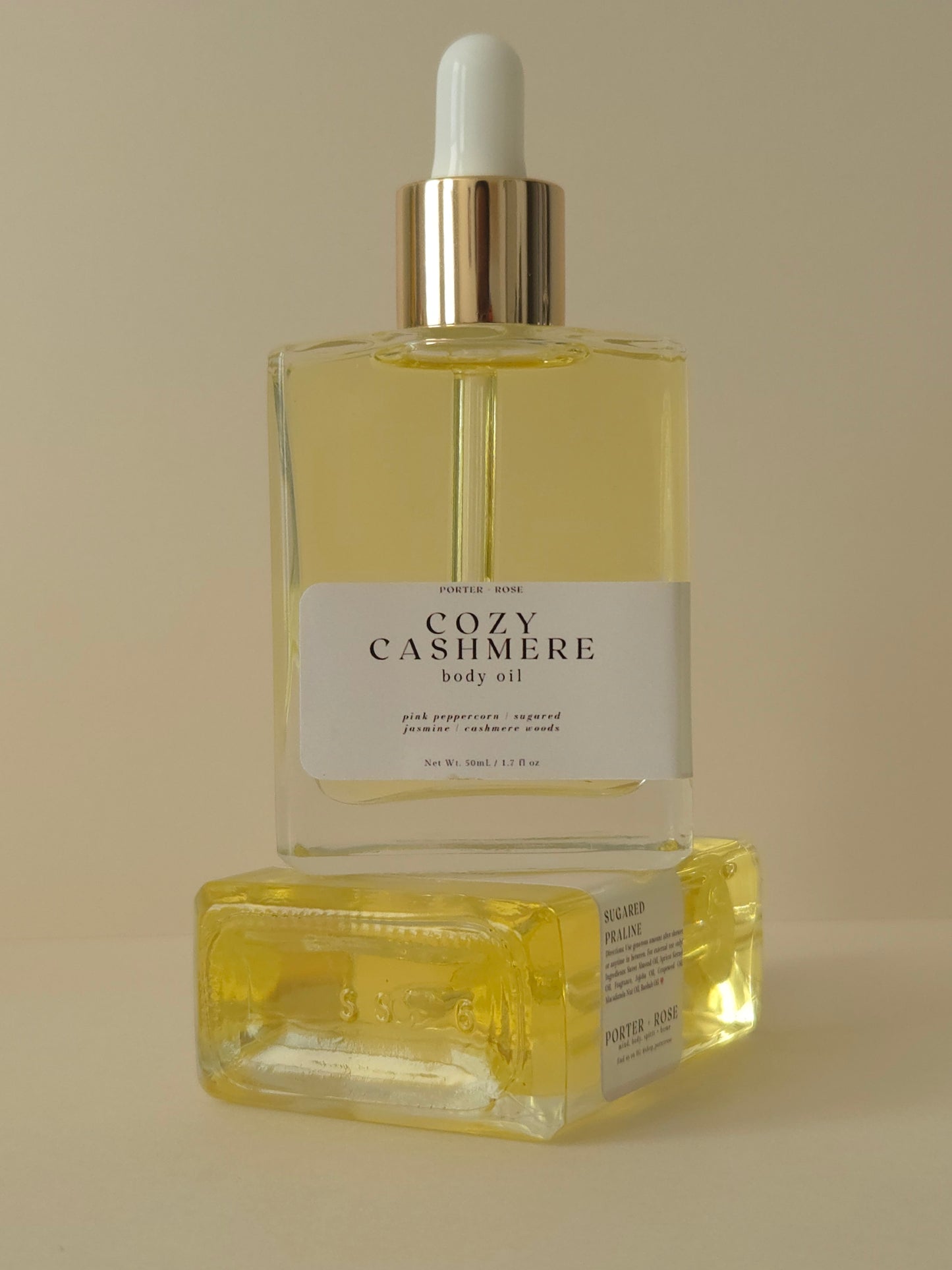 Two bottles of 'Cozy Cashmere' body oil on a beige background