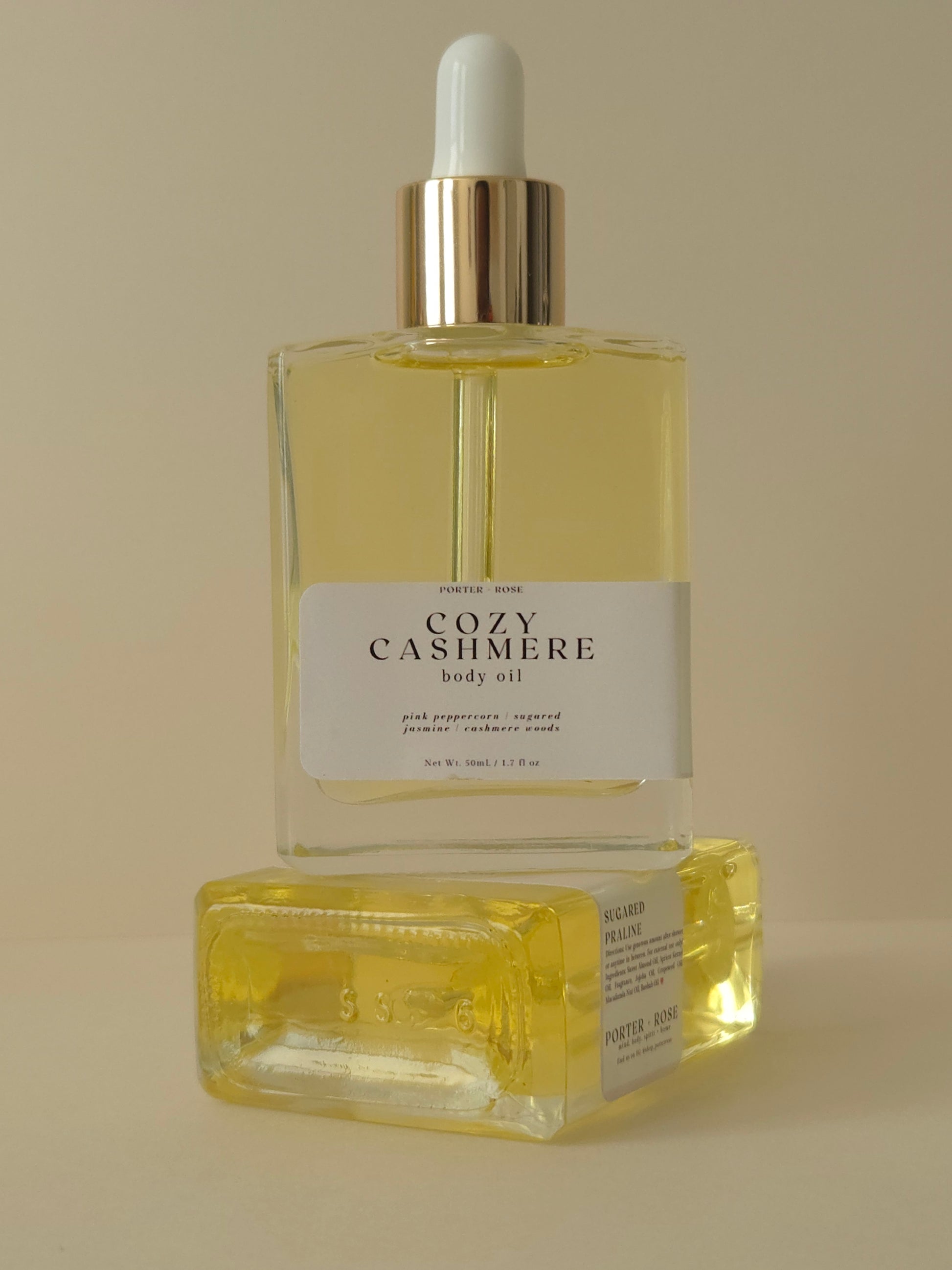 Two bottles of 'Cozy Cashmere' body oil on a beige background