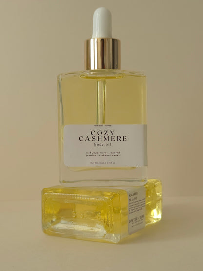 Two bottles of 'Cozy Cashmere' body oil on a beige background