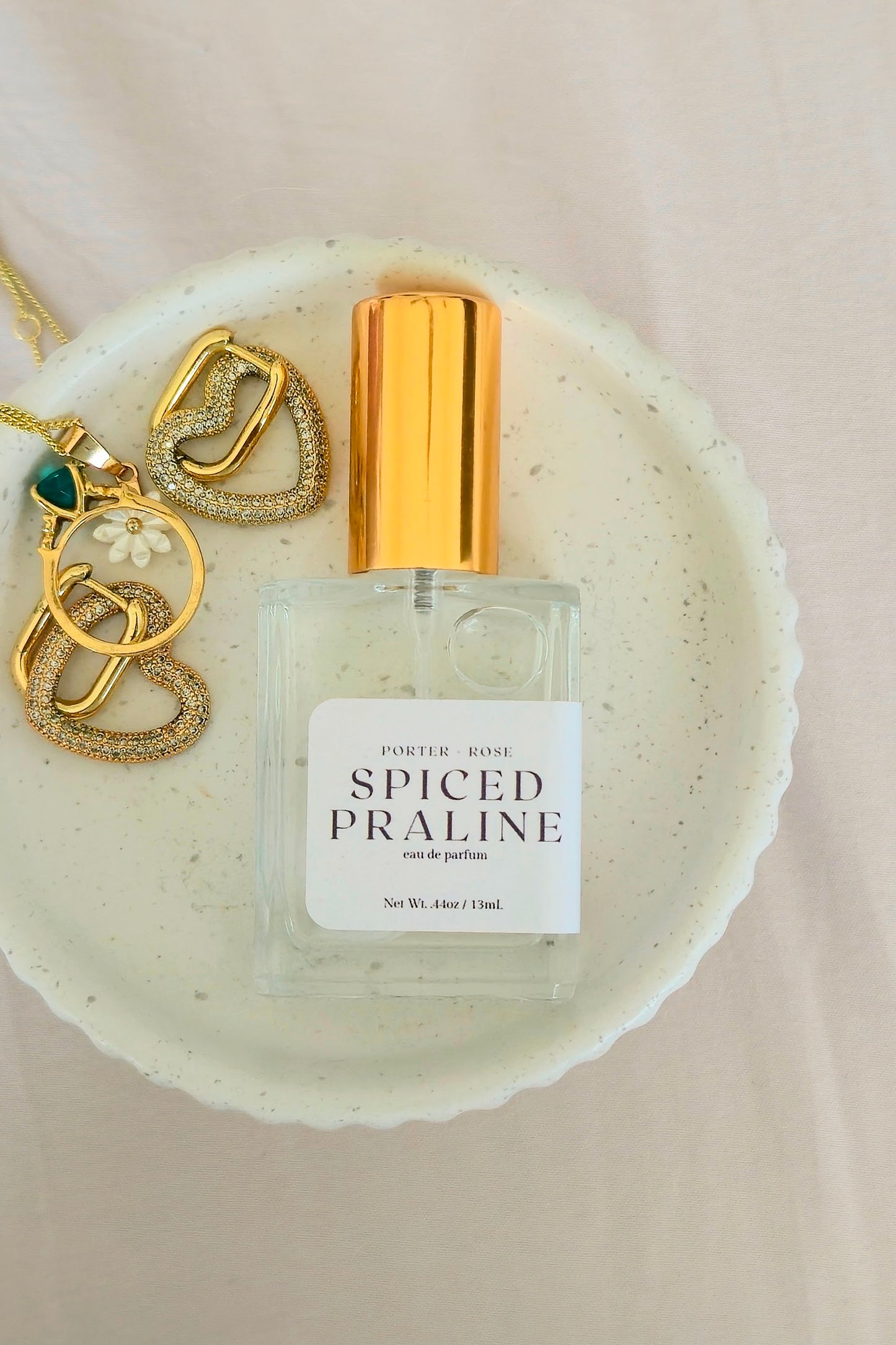 A bottle of Porter Rose Spiced Praline perfume with a gold cap and white label, styled on a speckled ceramic dish with gold jewelry against a soft beige fabric background.