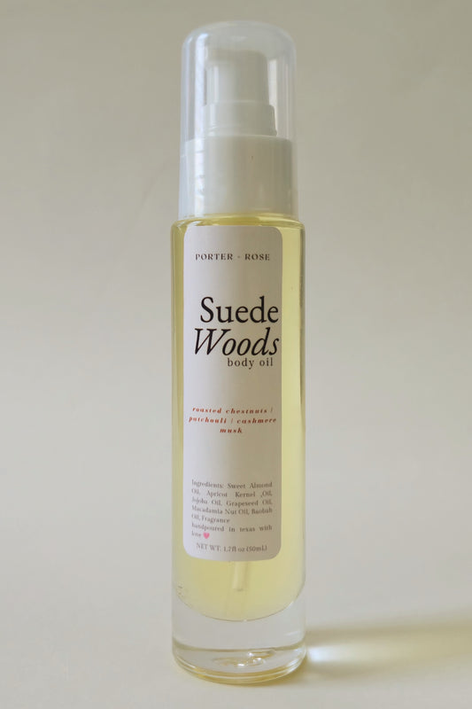 A bottle of Suede Woods body oil by Porter + Rose, featuring a clear cap and a white label with black text, containing a pale yellow oil, set against a plain light beige background.
