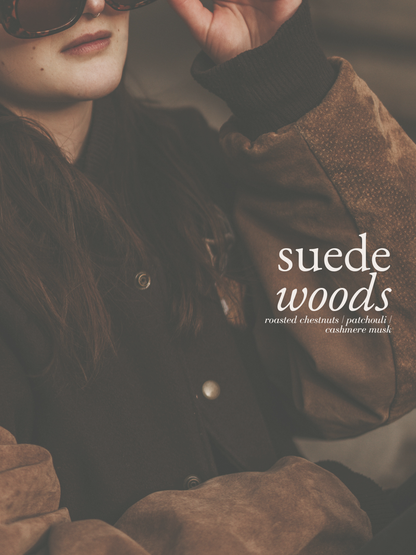 A close-up lifestyle shot of a model wearing a brown suede jacket with a black ribbed collar and cuffs, dark sunglasses, and long dark hair. The text 'suede woods' is overlaid on the right side.