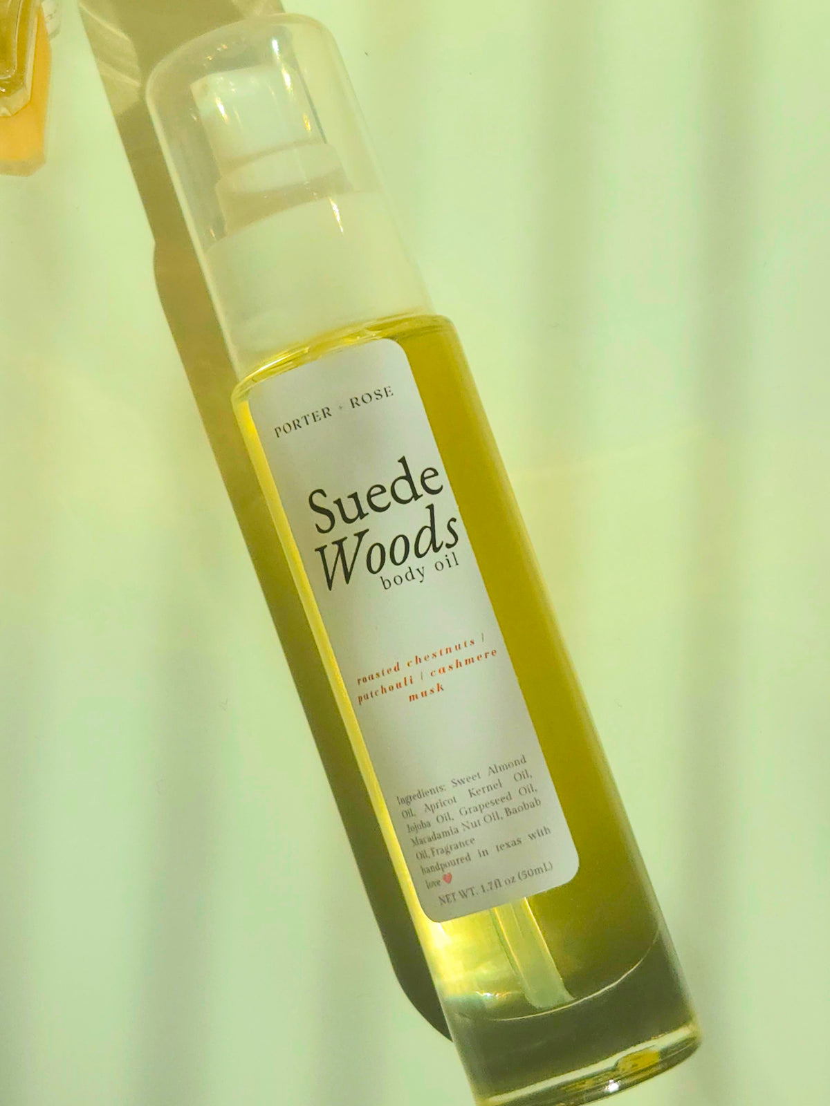 A bottle of Porter Rose Suede Woods Body Oil with a clear cap and a white label, filled with a yellow-green oil, set against a pale yellow background.