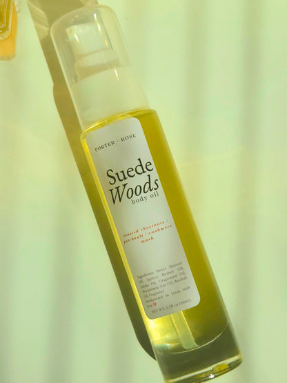 A bottle of Porter Rose Suede Woods Body Oil with a clear cap and a white label, filled with a yellow-green oil, set against a pale yellow background.