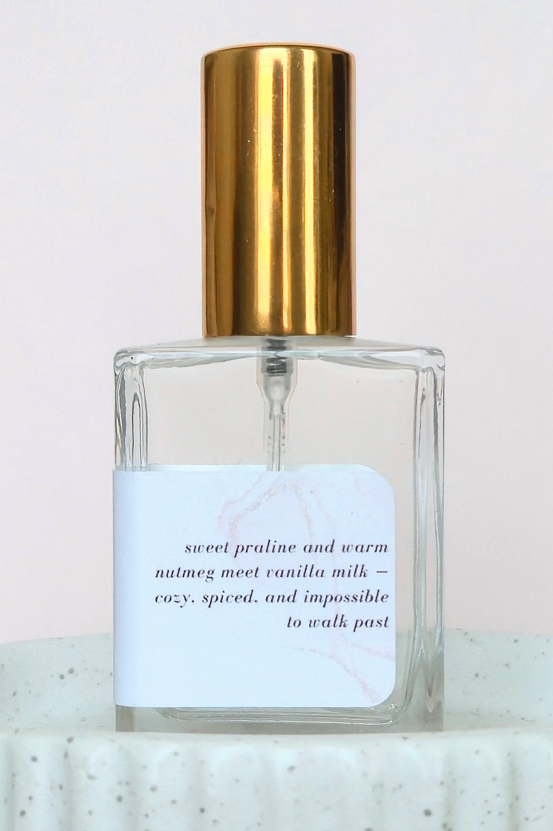 A clear glass perfume bottle with a shiny gold cylindrical cap and a white label featuring elegant black text, set against a soft light pink background.