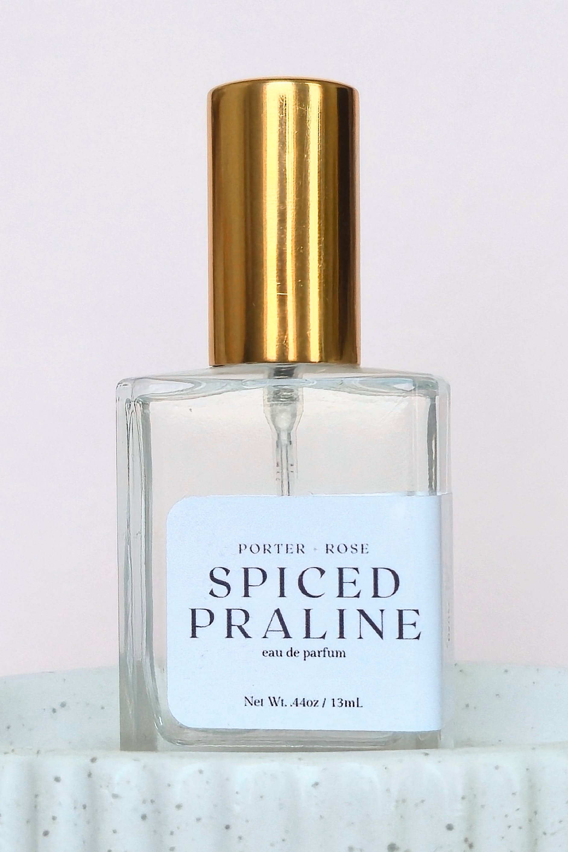 A clear glass perfume bottle labeled 'PORTER ROSE SPICED PRALINE eau de parfum' with a white label and a shiny gold cylindrical cap, set against a light pink background.