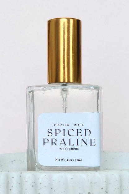 A clear glass perfume bottle labeled 'PORTER ROSE SPICED PRALINE eau de parfum' with a white label and a shiny gold cylindrical cap, set against a light pink background.