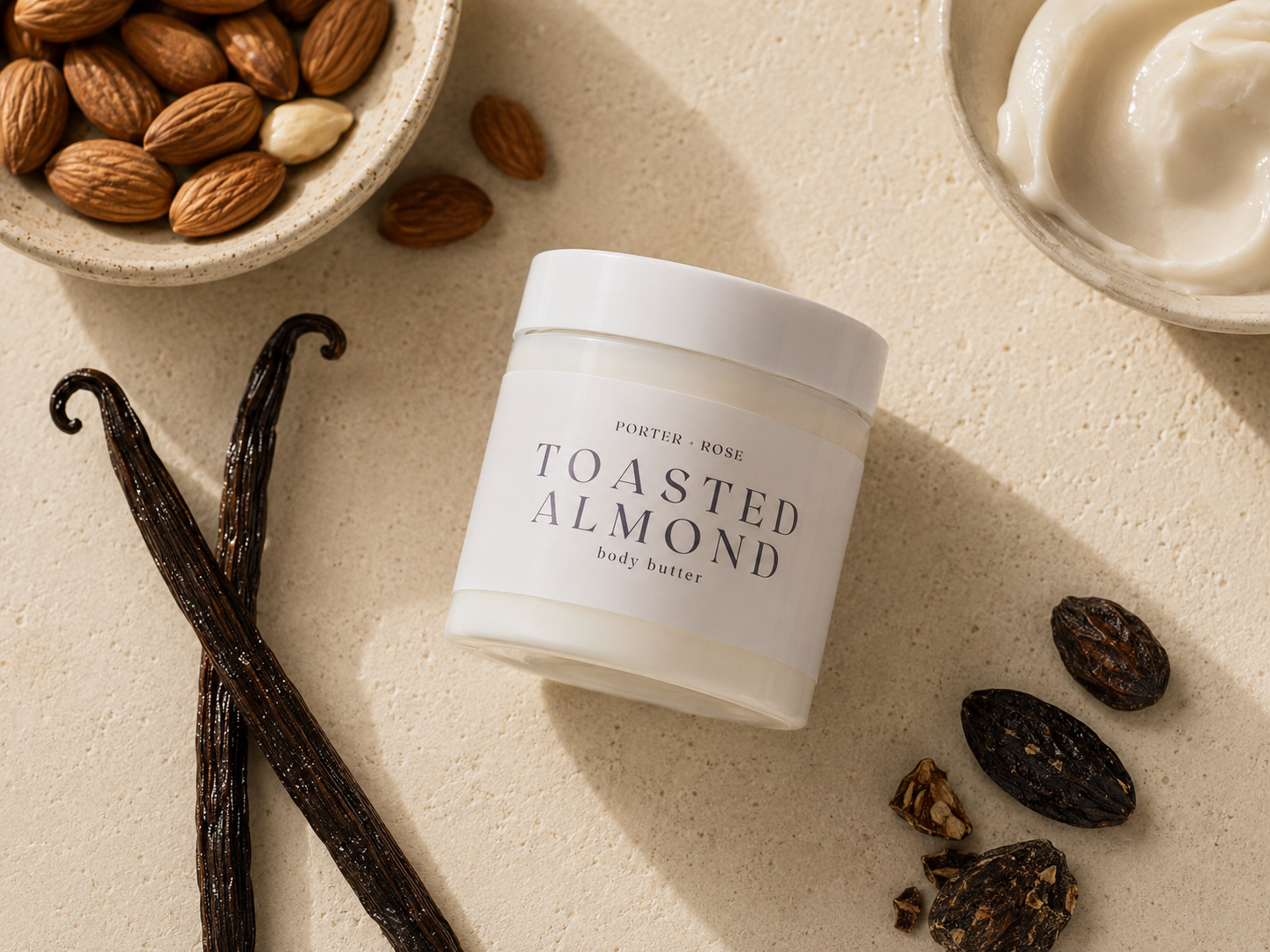 A jar of Porter Rose Toasted Almond Body Butter is displayed on a textured beige surface with its raw ingredients, including whole almonds in a bowl, vanilla beans, and dried almonds, arranged around it.