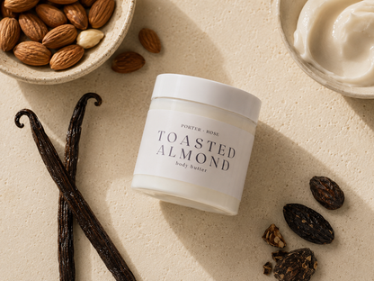 A jar of Porter Rose Toasted Almond Body Butter is displayed on a textured beige surface with its raw ingredients, including whole almonds in a bowl, vanilla beans, and dried almonds, arranged around it.