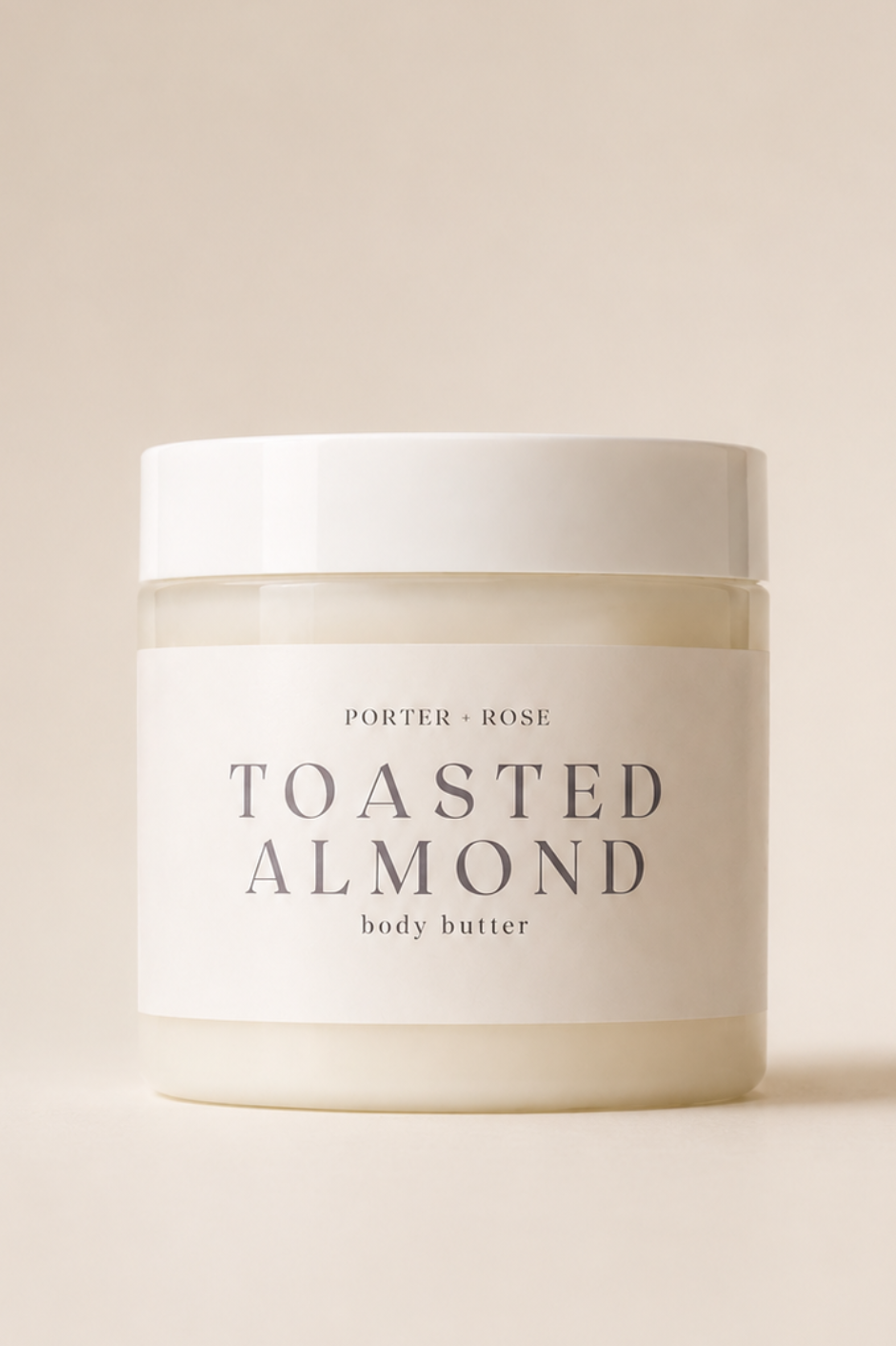 A jar of Porter + Rose Toasted Almond Body Butter on a warm beige background. The jar is white with a matching lid and a minimalist label featuring the product name in elegant serif lettering.