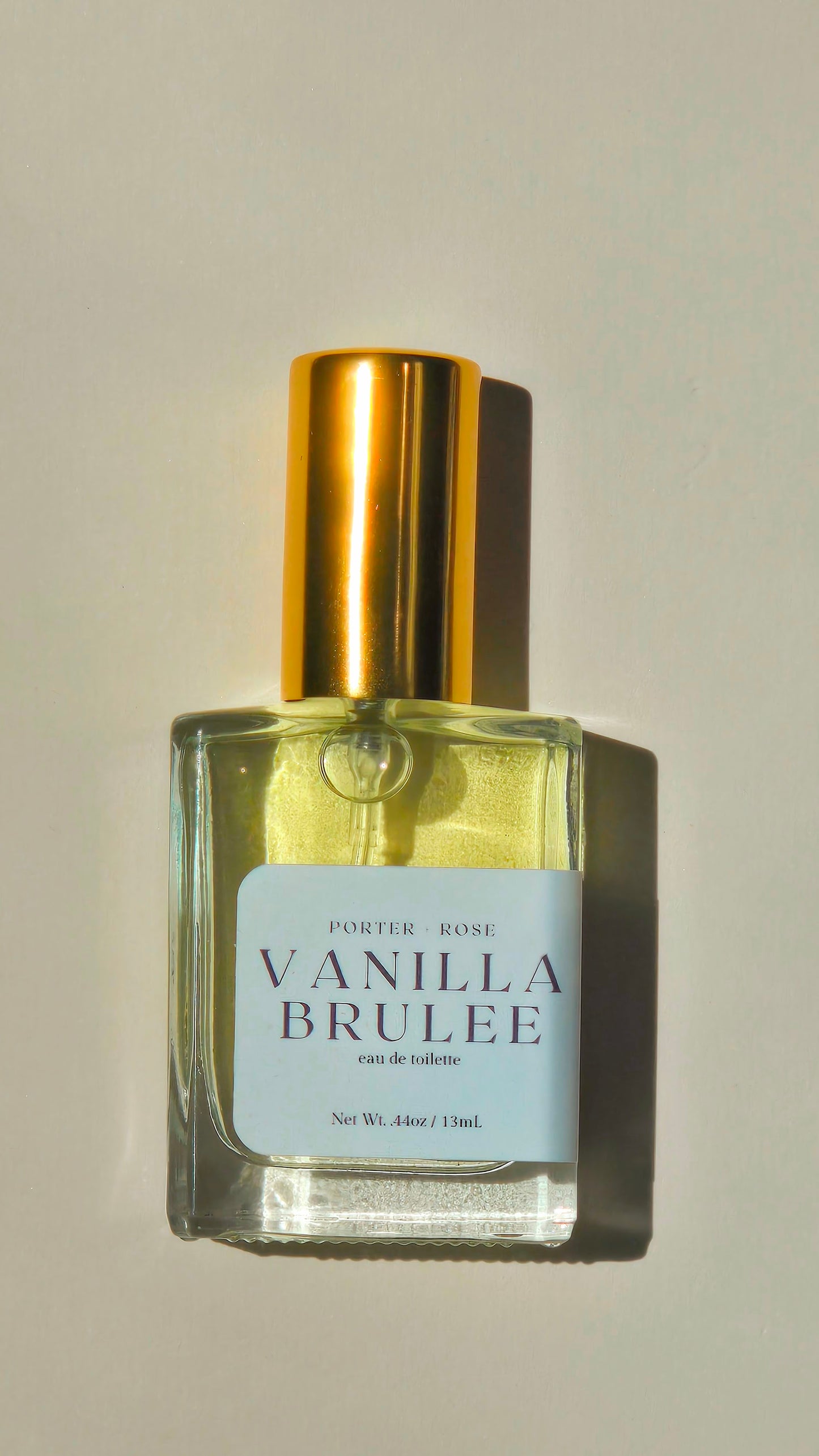 A bottle of Vanilla Brulee eau de toilette by Porter Rose, featuring a clear glass bottle with pale yellow liquid, a metallic gold cap, and a white label, set against a warm beige background with a soft shadow.