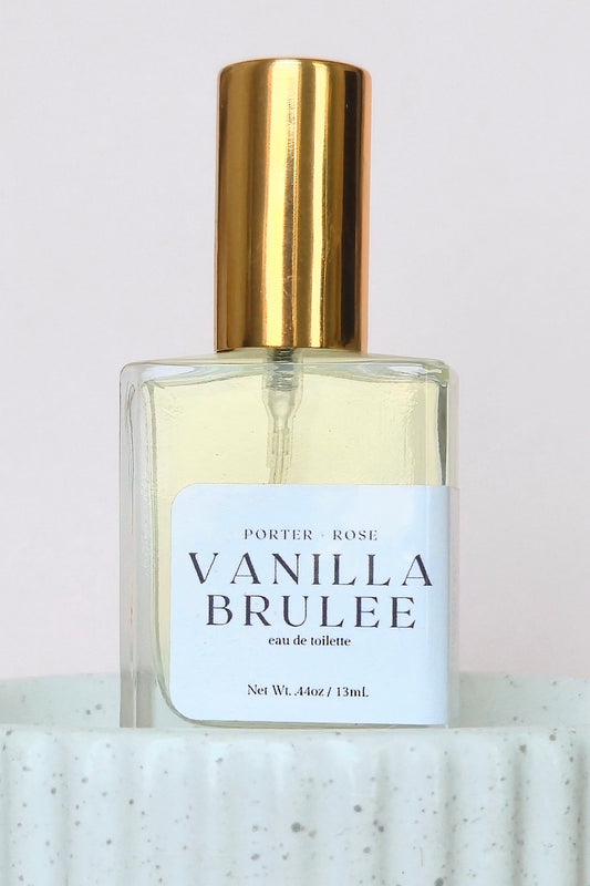 A bottle of Porter Rose Vanilla Brulee Eau de Toilette with a gold cap and a white label, displayed on a textured white ceramic surface against a light background.
