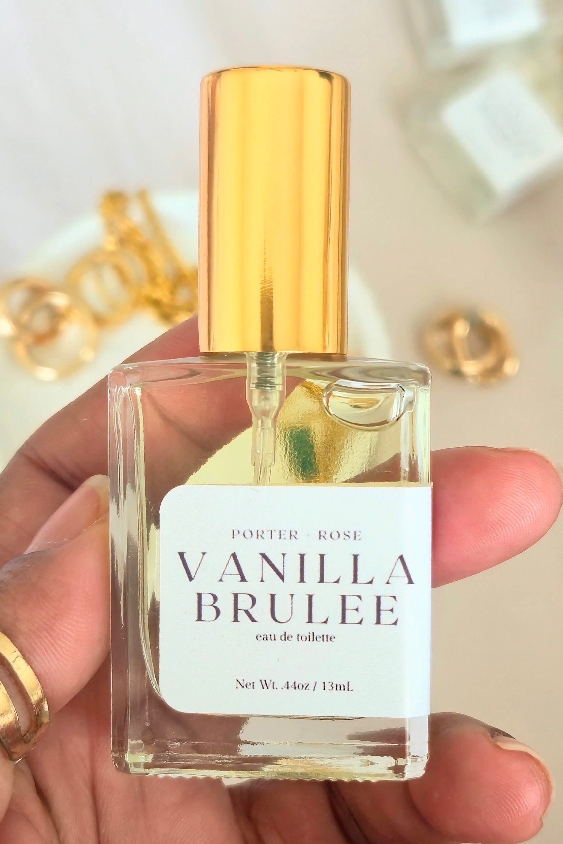 A hand holding a clear glass perfume bottle with a shiny gold cap and a white label that reads 'PORTER ROSE VANILLA BRULEE'. The background is a soft beige with blurred gold jewelry.