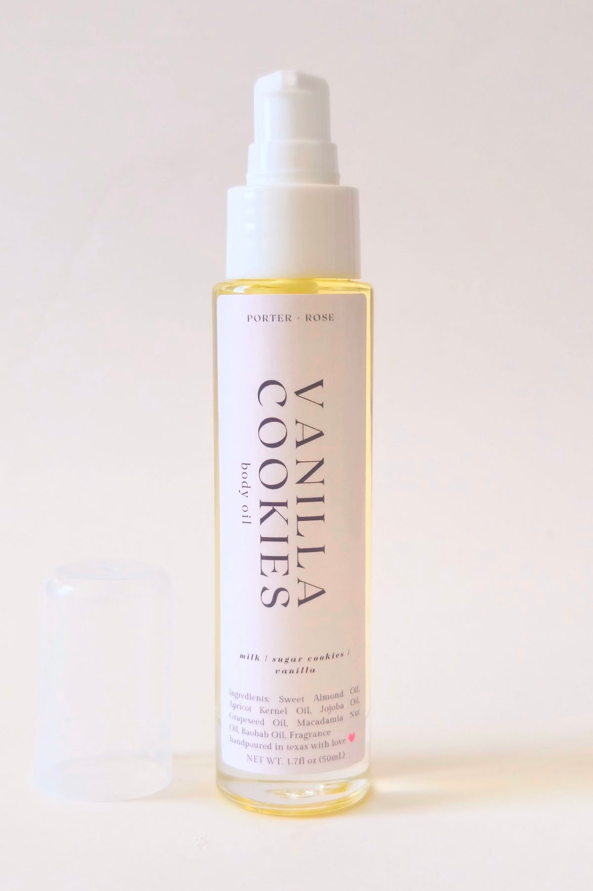 A bottle of Vanilla Cookies body oil with a white pump dispenser and a translucent cap resting beside it on a clean, light beige background.