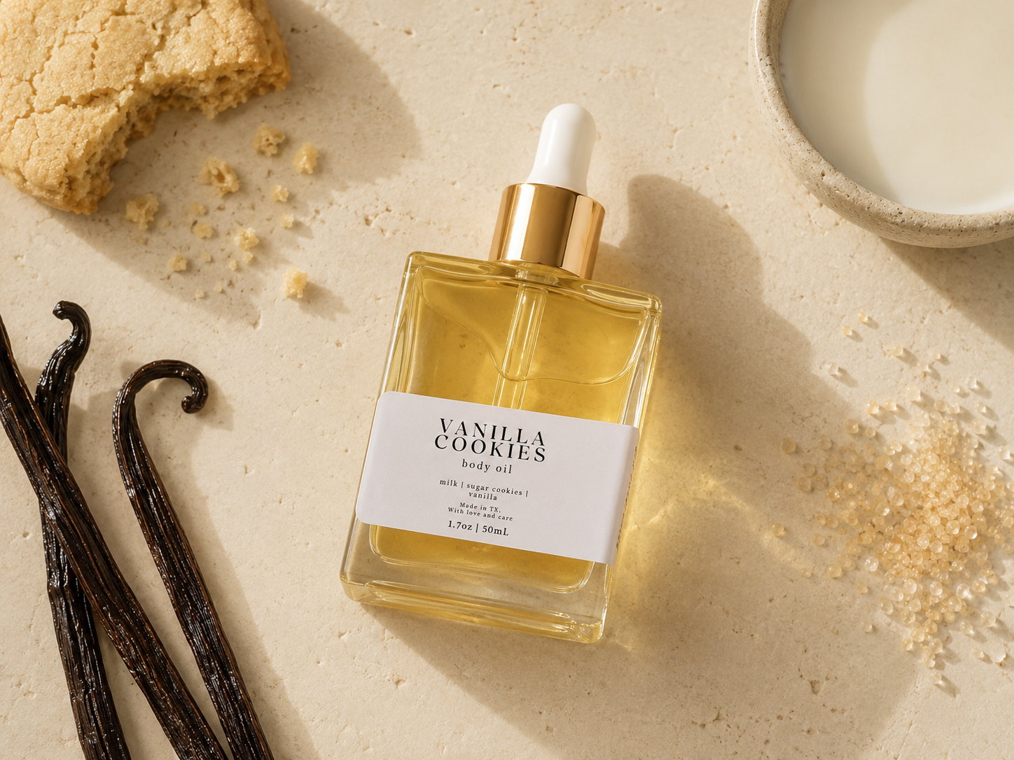 A bottle of Vanilla Cookies body oil with a white label and gold dropper cap, styled on a beige surface with a cookie, vanilla beans, sugar, and a bowl of milk.