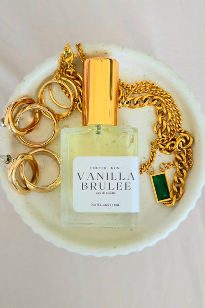 A bottle of Porter Rose Vanilla Brulee Eau de Parfum is displayed on a white ceramic dish with gold jewelry props, including rings and a chain with a green pendant, on a light beige fabric background.